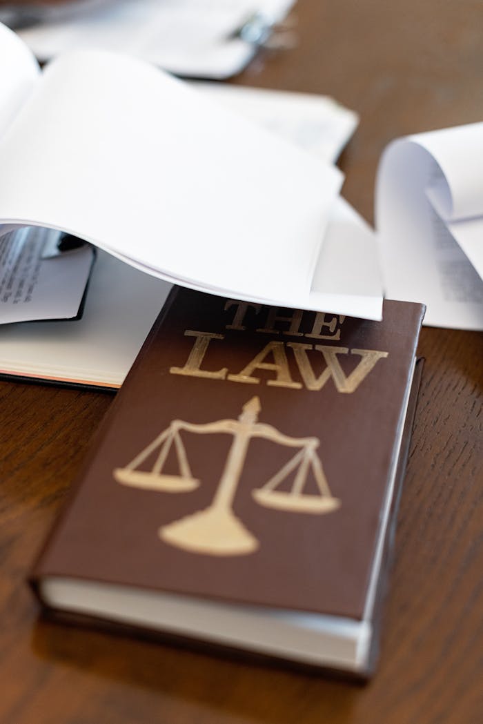 hero-img-01 Close-up of a law book with scales icon on a wooden desk covered in papers.
